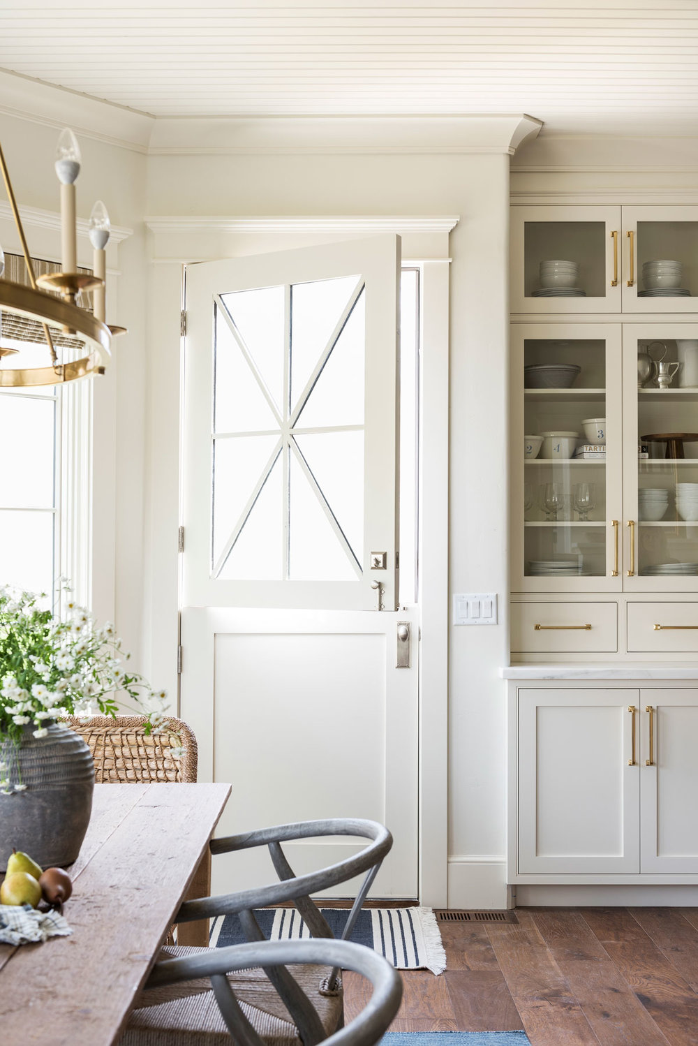 Charming neutral kitchen and dining space featuring a white Dutch door with geometric window panes, antique hardwood floors, and built-in white cabinetry with warm brass hardware