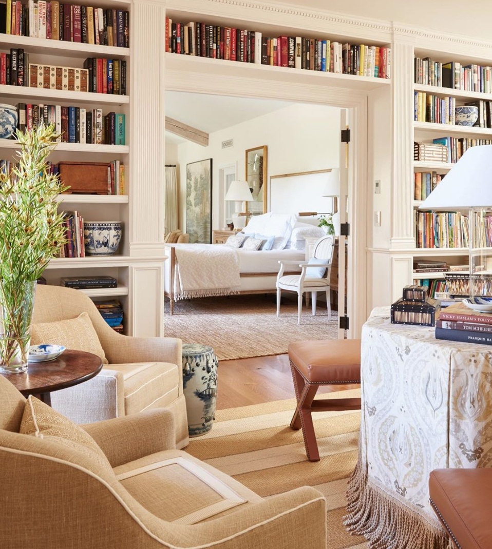 Neutral transitional home library and sitting room featuring custom floor-to-ceiling cream bookshelves filled with books. In the foreground, two tan upholstered club chairs with white piping rest on a striped neutral area rug over hardwood floors. A wide architectural doorway provides a view into a serene white bedroom with a canopy bed
