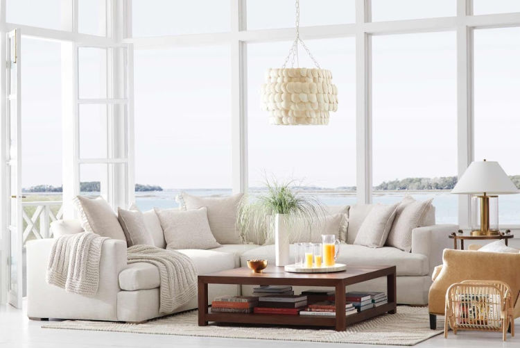 A sunlit coastal living room with a white sectional couch, a square dark wood coffee table stacked with books, and an elegant capiz shell chandelier hanging from the ceiling. Large panoramic windows reveal a serene bay or marsh view under a bright sky