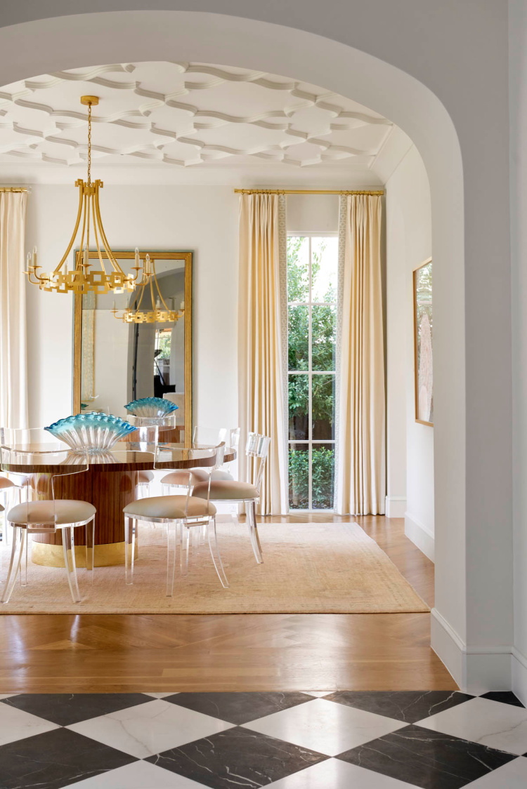 Luxurious dining room viewed through an archway, featuring a detailed geometric fretwork ceiling, a round walnut table with acrylic chairs, a large gold chandelier, and a black and white checkerboard marble foyer floor