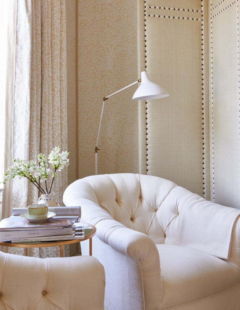 Warm monochromatic neutral interior design featuring an upholstered ivory tufted club chair and a cream throw blanket. A modern white adjustable floor lamp highlights a textured grasscloth wall panel with vertical nailhead trim. The background displays elegant soft gold damask wallpaper, a small brass end table with magazines and a white flower vase, and matching patterned linen curtains